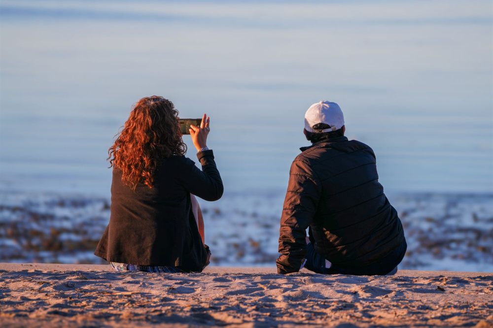 Couple sitting on the sand photographing the sea at sunset